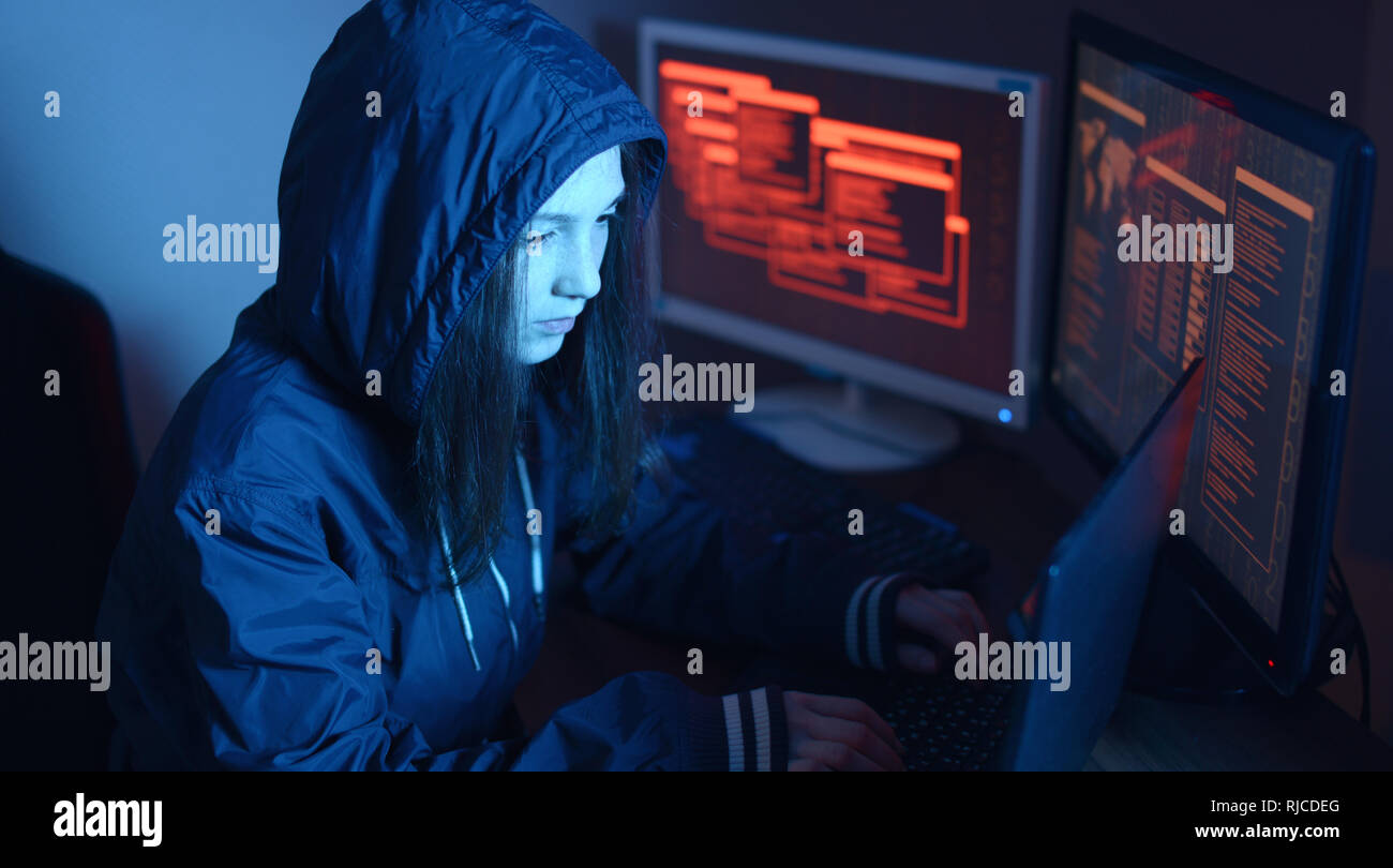 Ragazza hacker in una cappa digitando il codice di un programma mentre commette un crimine cibernetico hacking un firewall di sistema sullo sfondo delle schermate in scuro sotto il neon Foto Stock