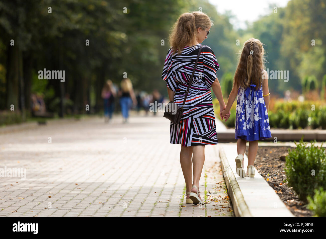 Vista posteriore del giovane biondo con i capelli lunghi slim donna attraente ed un bambino piccolo ragazza in abiti alla moda a piedi tenendo le mani da sunny summer park alle Foto Stock