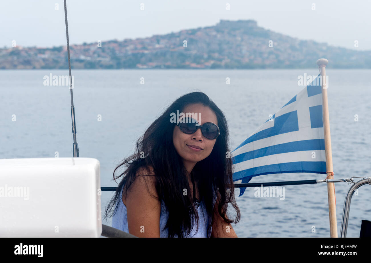 Donna in occhiali da sole seduto in cabina di pilotaggio di barca a vela  con bandiera greca e la città di MOLIVOS, LESBO Foto stock - Alamy