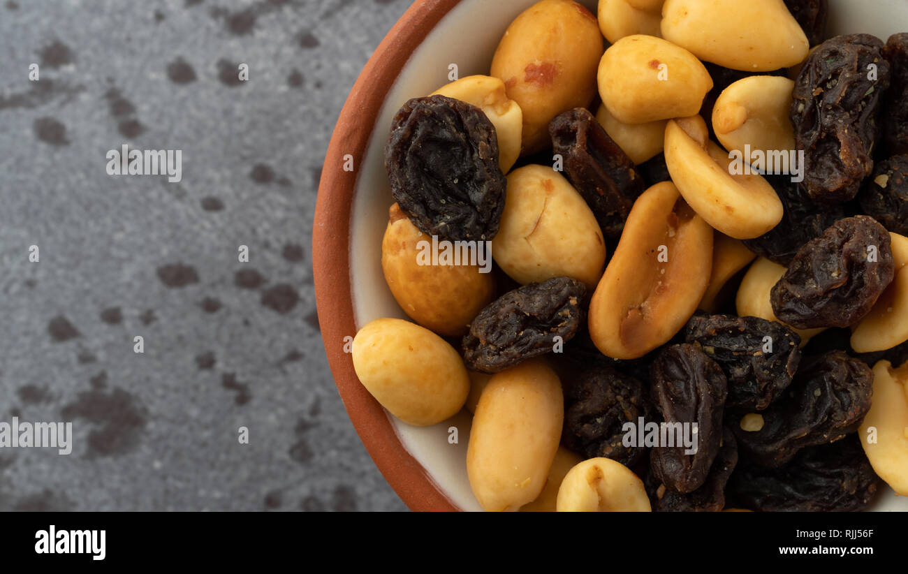 Top vista ravvicinata di una piccola ciotola riempita con arachidi e uva passa su sfondo grigio illuminato con luce naturale. Foto Stock