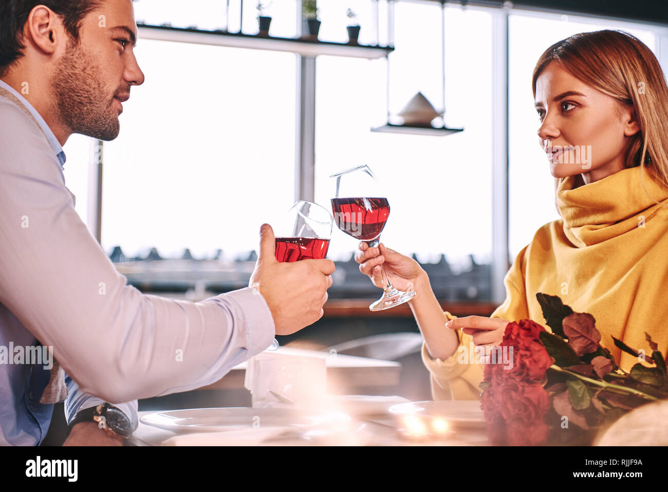 Momenti solenni. Giovane Azienda vino rosso in bicchieri nel ristorante. Rose rosse e candele sono giacenti sul tavolo vicino al vino rosso. Giovane donna in musta Foto Stock