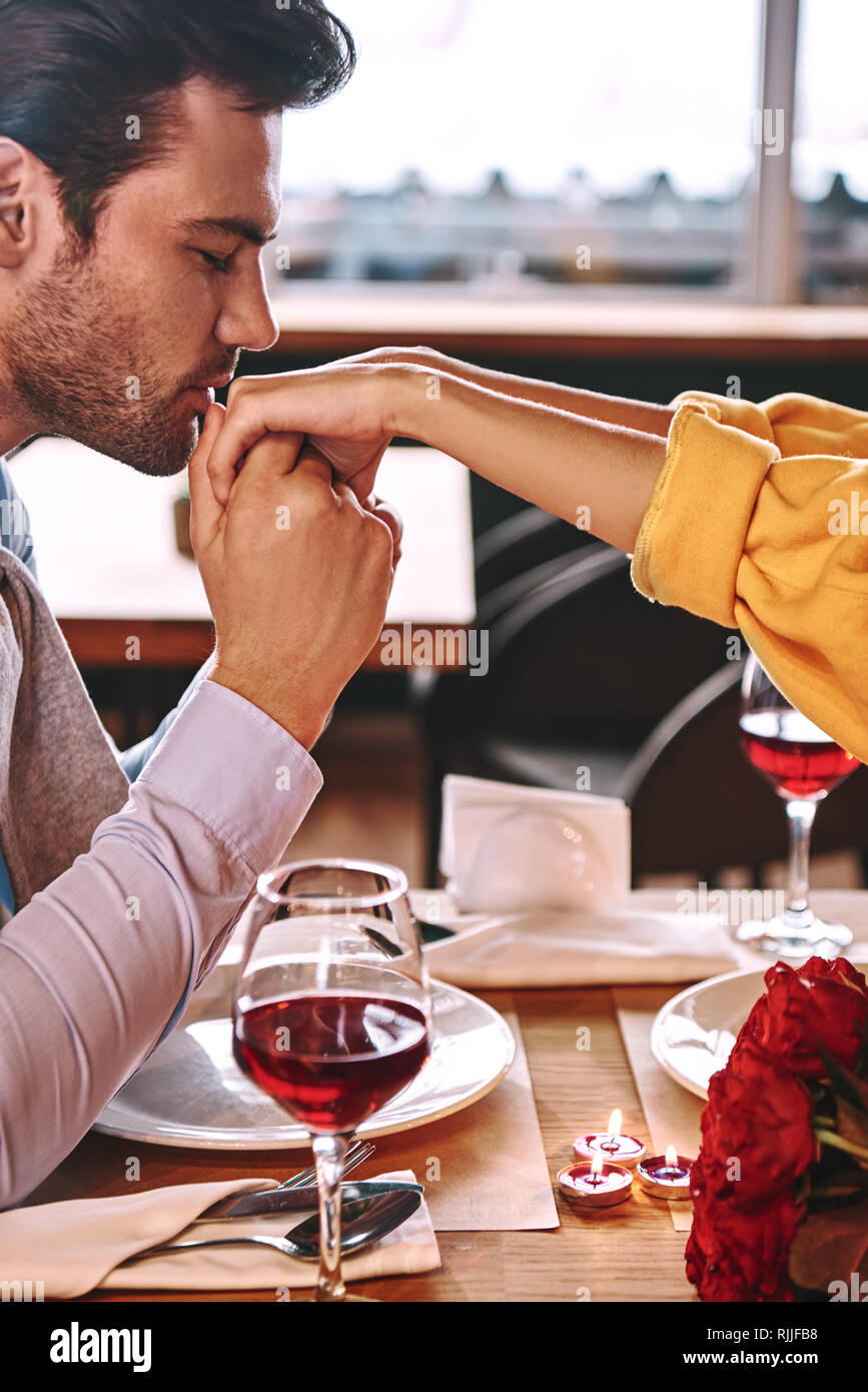 Proposta romantica. Giovane uomo bacia le mani della sua fidanzata nel ristorante. Rose rosse e candele sono giacenti sul tavolo vicino al vino rosso. Pho ritagliata Foto Stock