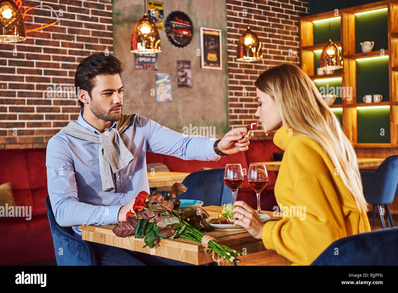 Cena romantica. Uomo in camicia blu sfamare la sua bella ragazza bionda nel ristorante. Rose rosse sono sdraiato sul tavolo. Giovane donna bionda senape in sw Foto Stock