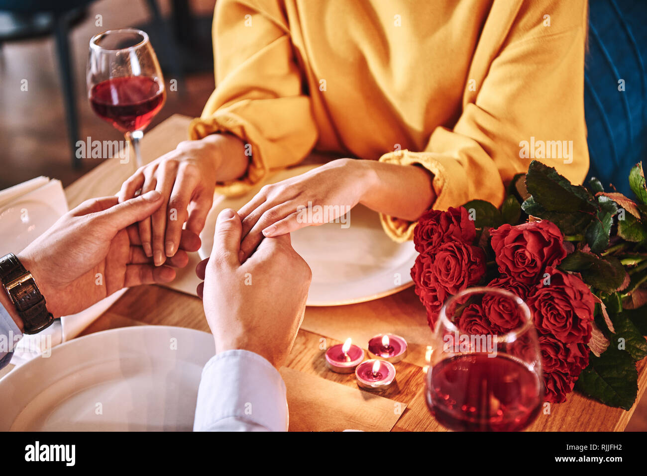Momenti romantici. Giovane uomo tenendo le mani della sua fidanzata nel ristorante. Rose rosse e candele sono giacenti sul tavolo vicino al vino rosso. Giovane donna Foto Stock