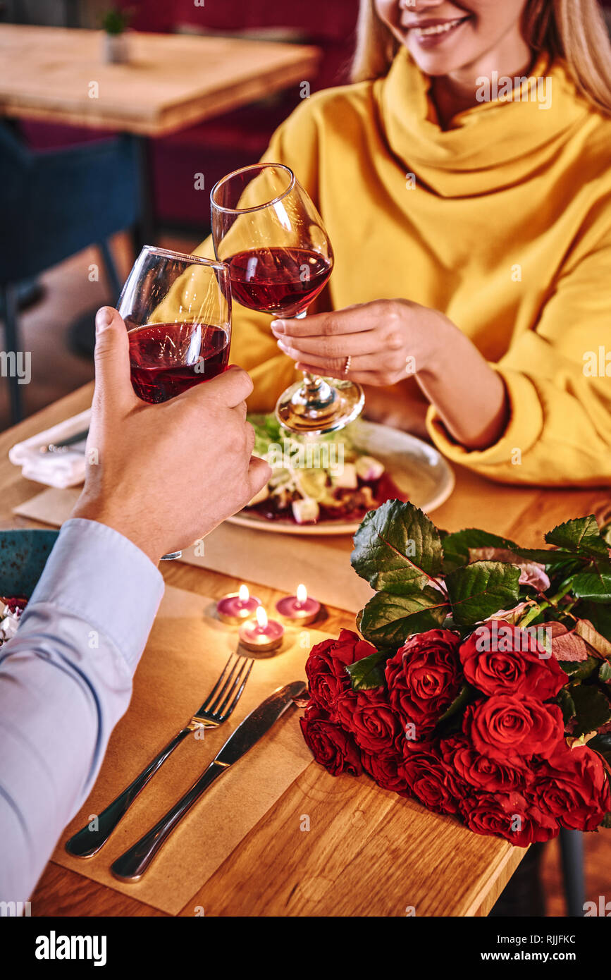 Il tifo per la felicità. Close-up del giovane azienda vino rosso in bicchieri nel ristorante. Rose rosse e candele sono giacenti sul tavolo vicino al vino rosso. Yo Foto Stock