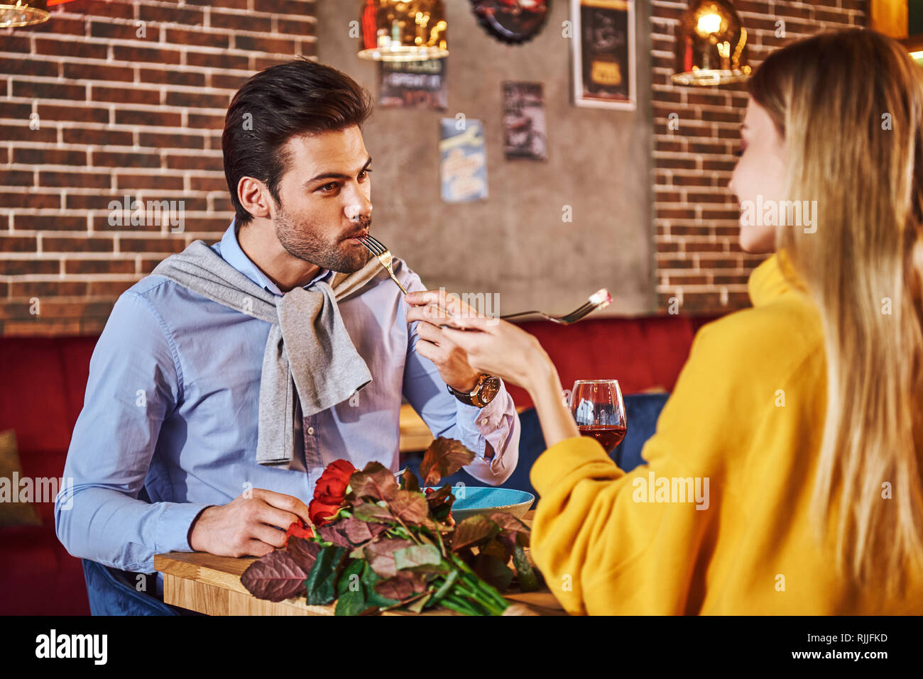 Necessità di alimentare il suo. Uomo in camicia blu mangiare e parlare con la fidanzata a cena romantica. Rose rosse sono sdraiato sul tavolo. Giovane donna bionda in Foto Stock