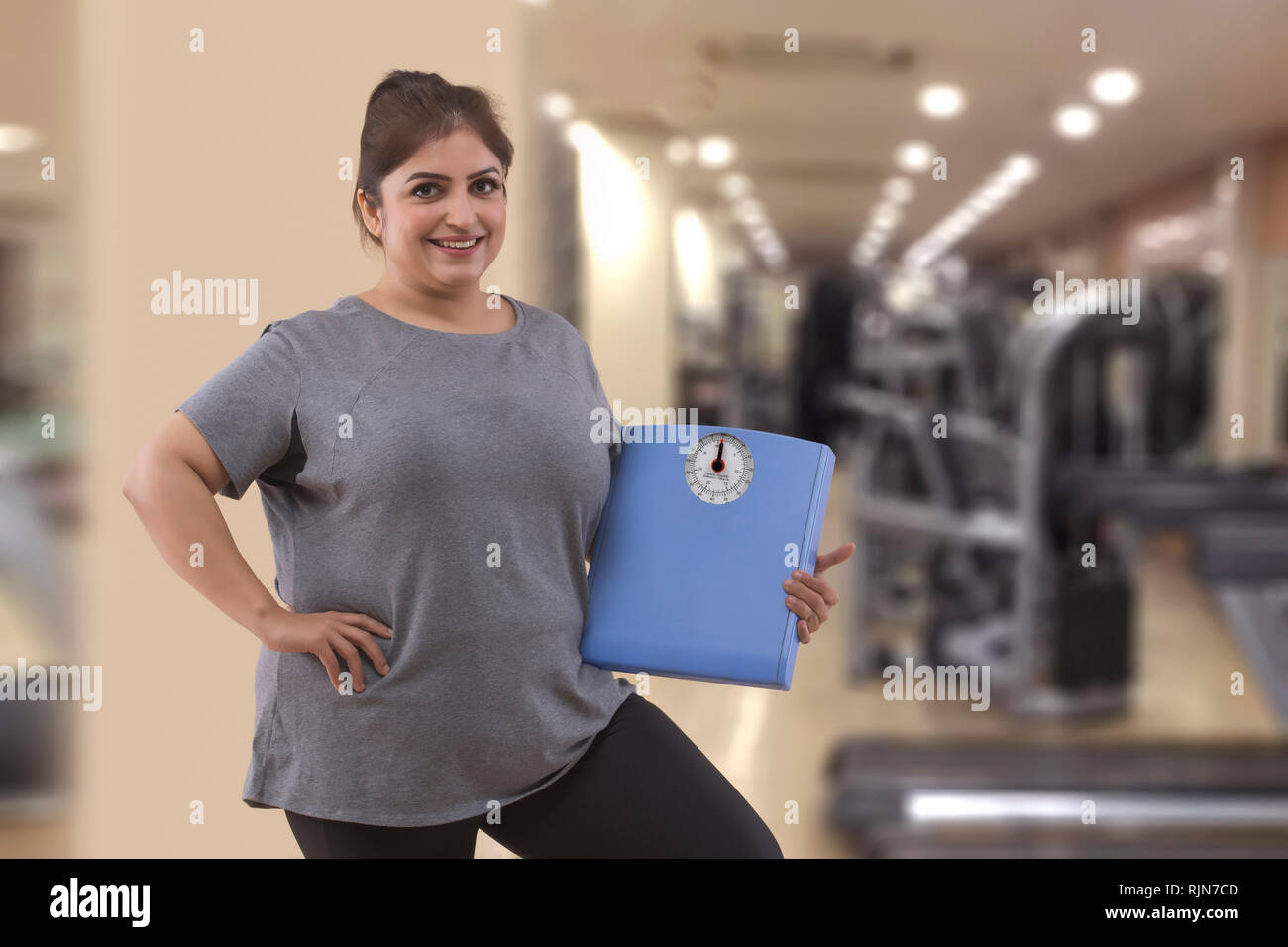 Sorridente donna grassa in una palestra in possesso di una bilancia pesapersone Foto Stock