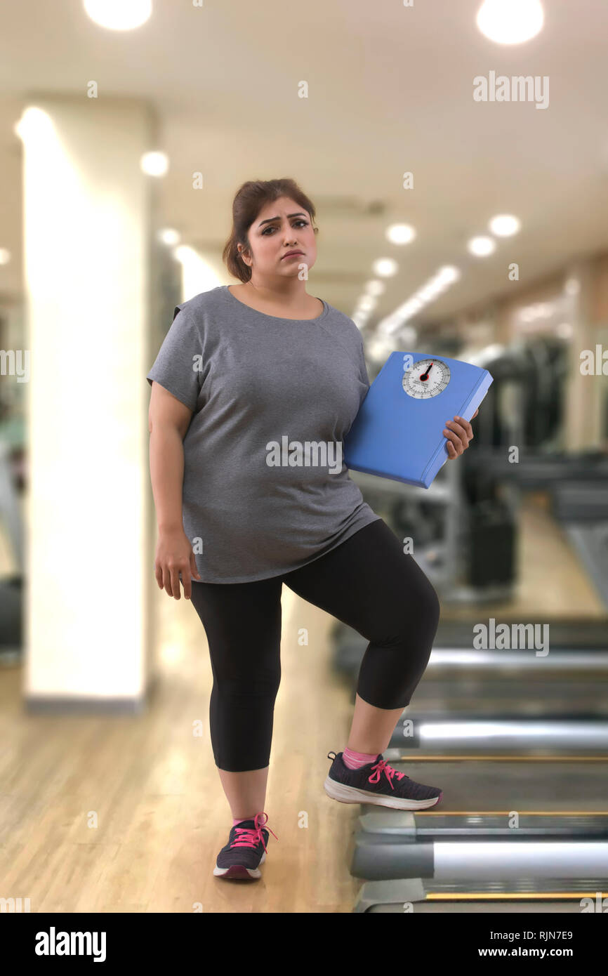 Donna grassa in una palestra in possesso di una bilancia pesapersone Foto Stock
