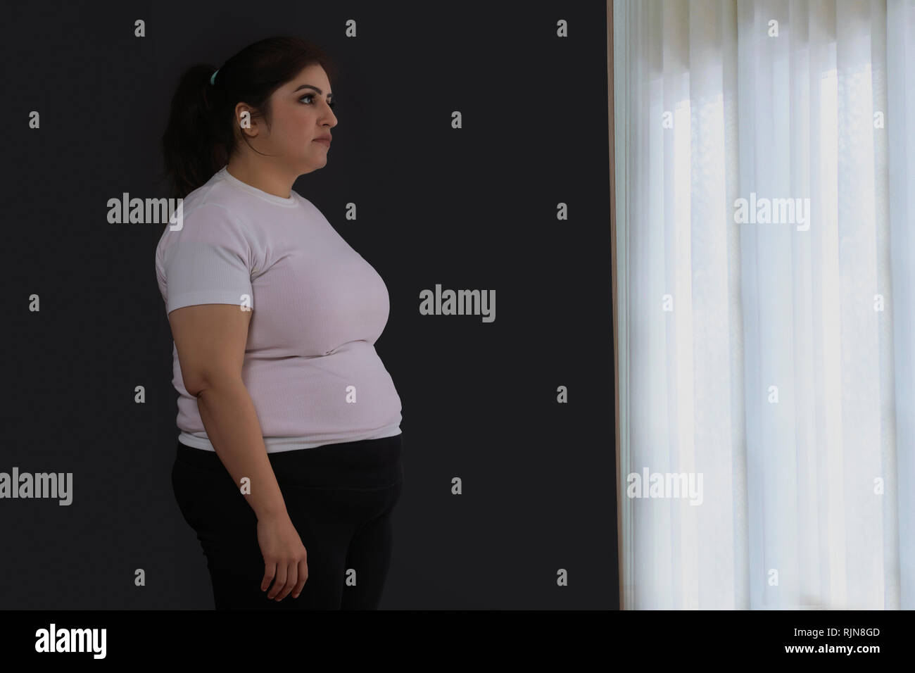 Triste donna obesa guarda stando in piedi dalla finestra Foto Stock