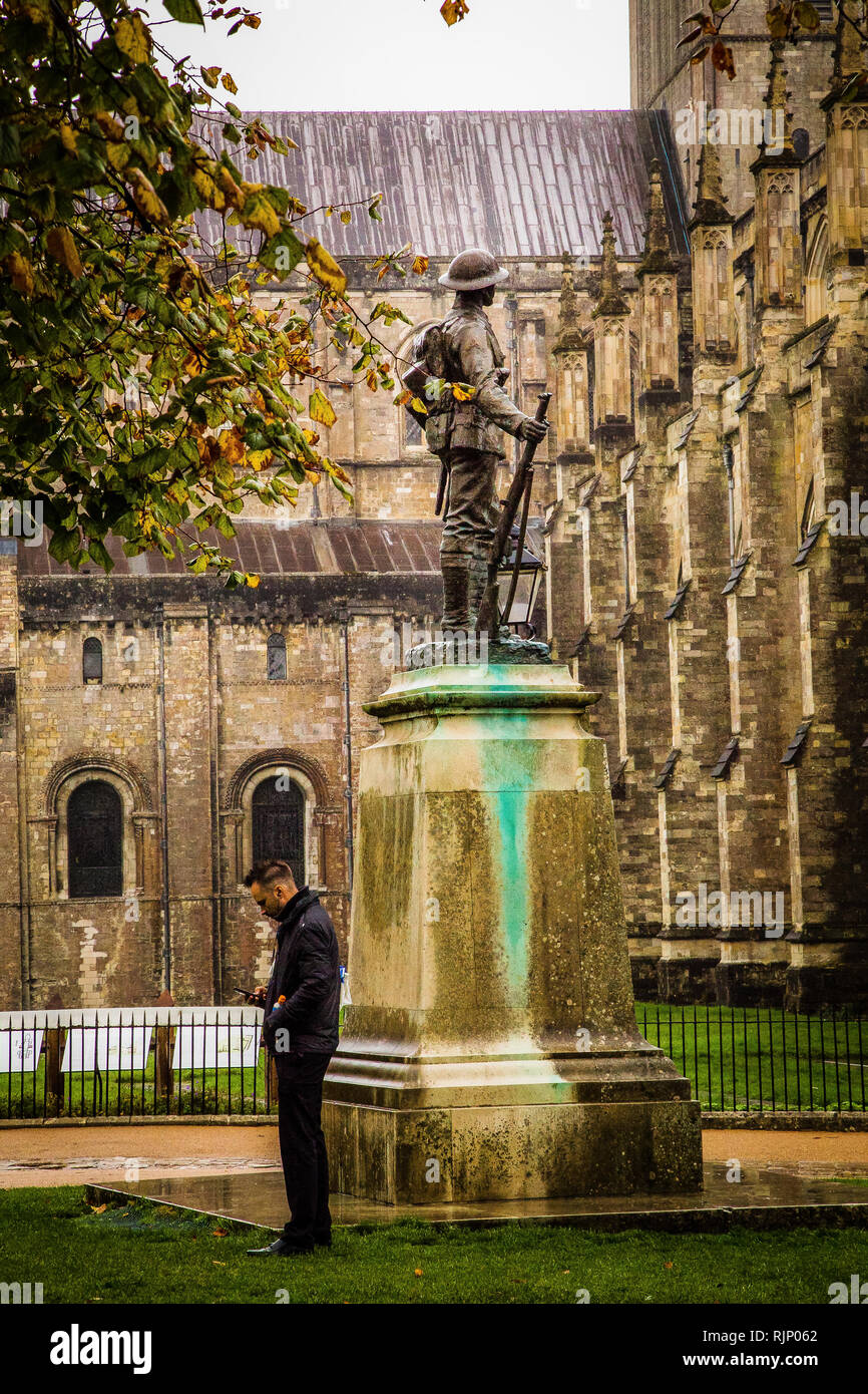 Winchester, Hampshire. Uomo in piedi da un memoriale di guerra alla ricerca di un telefono cellulare con alberi e la Cattedrale di Winchester anche in vista. Foto Stock