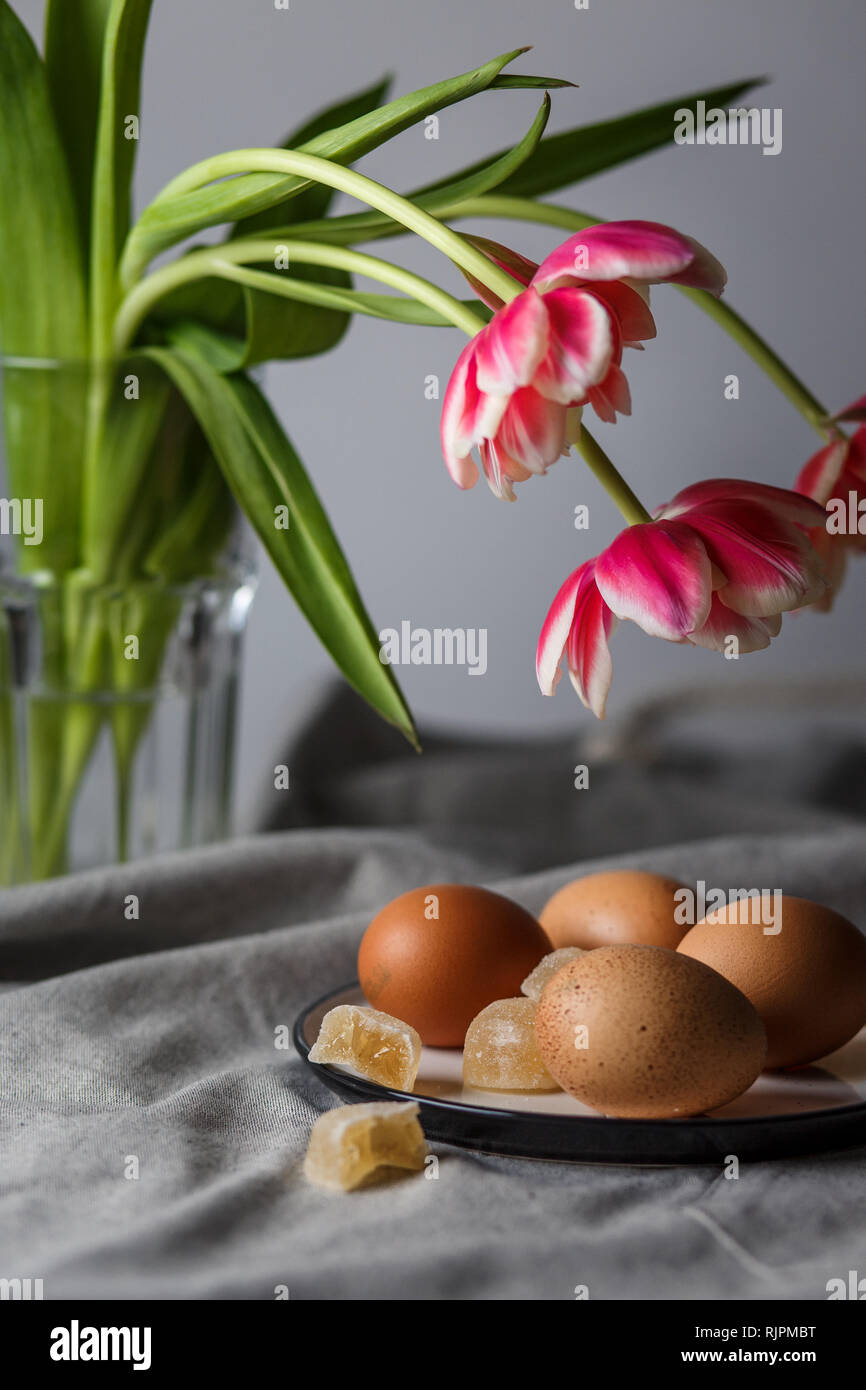 Bollite le uova di colore marrone su una piastra vicino ai tulipani in un vetro trasparente cup su sfondo grigio in una luce ovattato e in soft focus Foto Stock