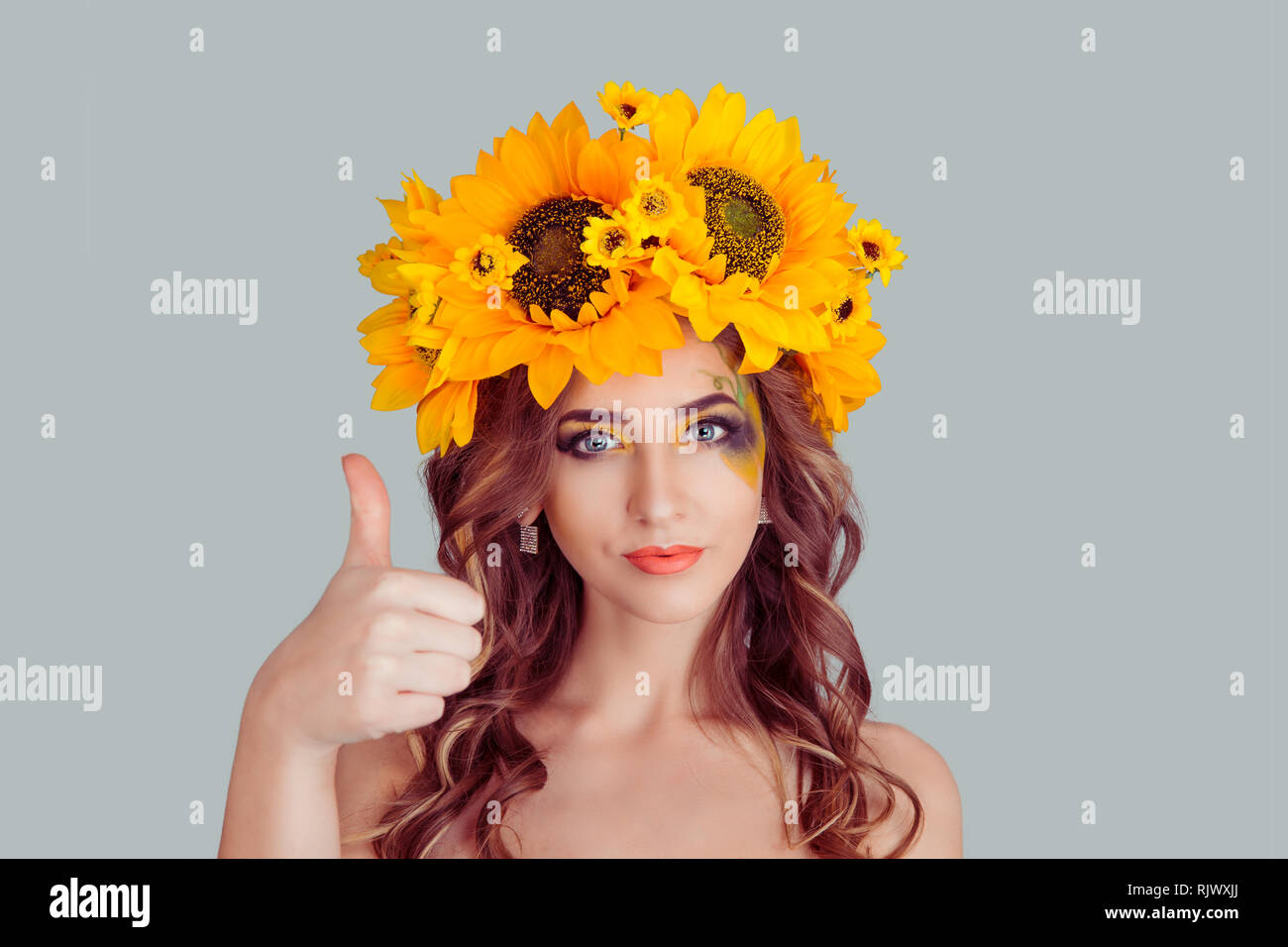 Felice giovane donna con fascia floreale che mostra i pollici come segno di mano. Ragazza con corona da girasoli sulla testa isolata su uno sfondo grigio con cop Foto Stock
