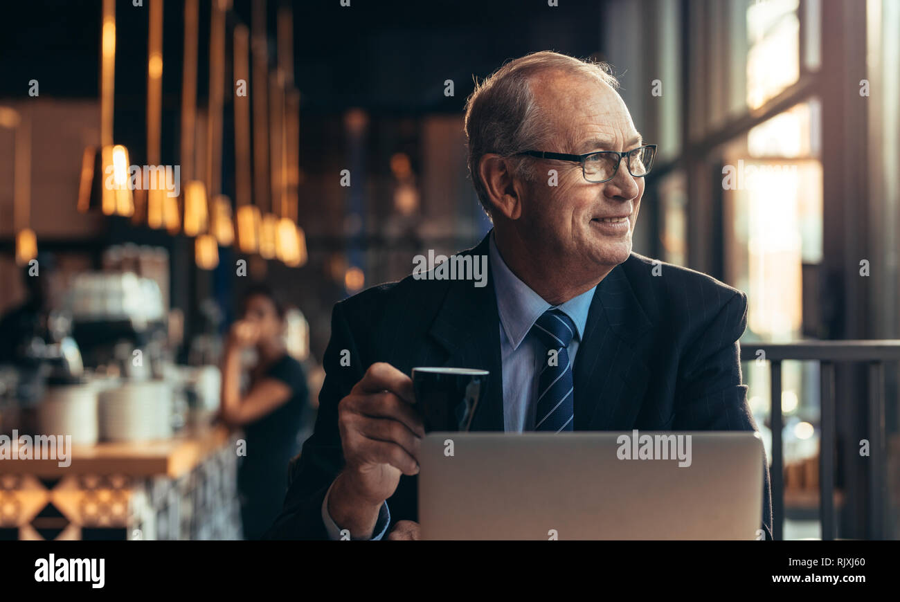Rilassata imprenditore senior seduto al caffè con caffè in mano guardando lontano sorridente. Maschio maturo imprenditore rilassante presso la caffetteria con il computer portatile in f Foto Stock