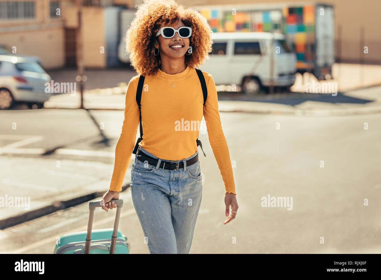 Donna afro american viaggiatore in vacanza a piedi intorno alla città tirando la sua valigia trolley. Sorridente turista donna che indossa gli occhiali da sole e uno zaino wa Foto Stock