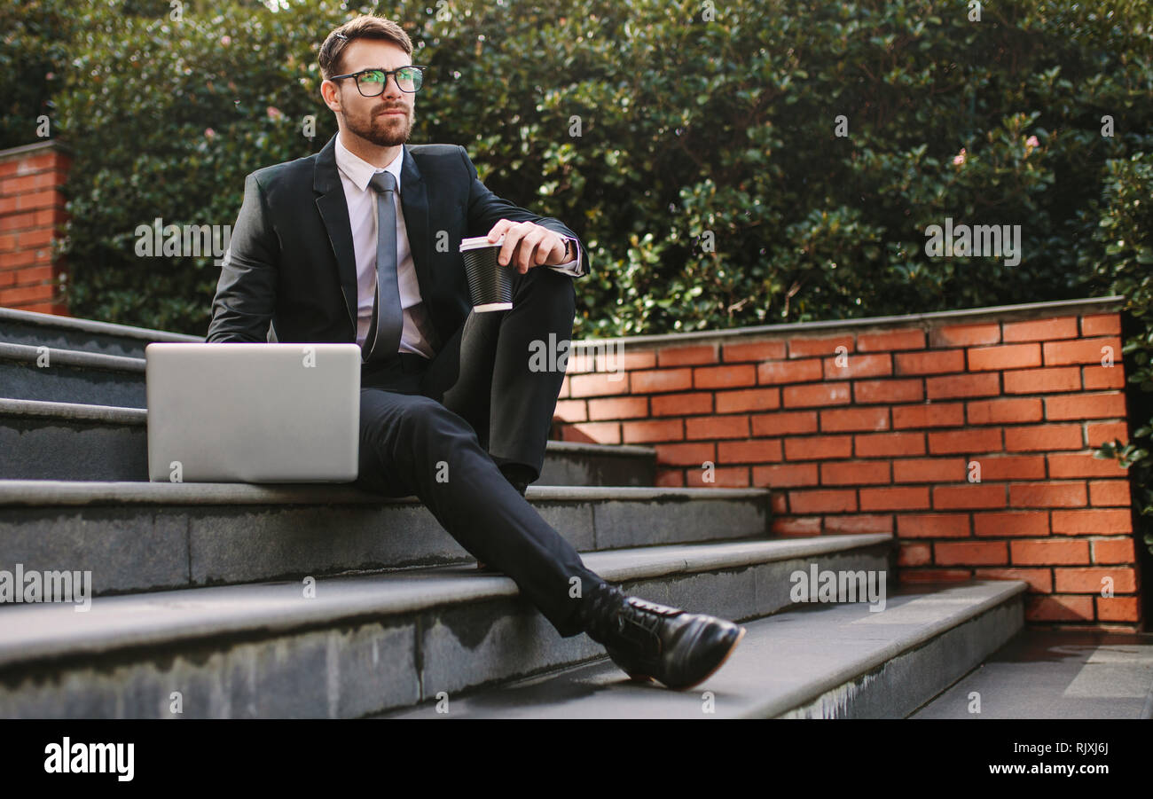 uomo d'affari che si rilassa sui gradini con un computer portatile e un caffè che si affacciano lontano. Imprenditore maschile che si rilassa all'aperto con caffè e laptop. Foto Stock