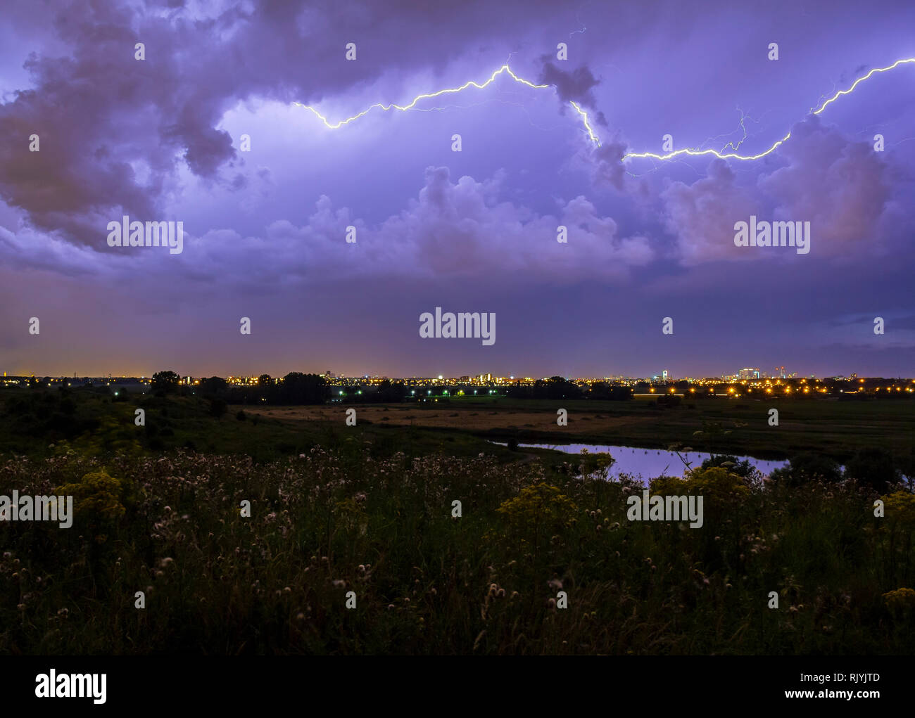 Intracloud fulmine IC thunderbolt colpisce di notte al di sopra di una città. Drammatica cloudscape temporale, colline con fiori sul primo piano, città sul Foto Stock