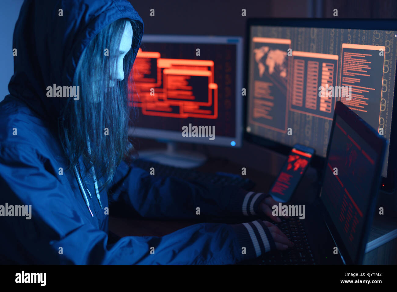 Ragazza hacker in una cappa digitando il codice di un programma mentre commette un crimine cibernetico hacking un firewall di sistema sullo sfondo delle schermate in scuro sotto il neon Foto Stock