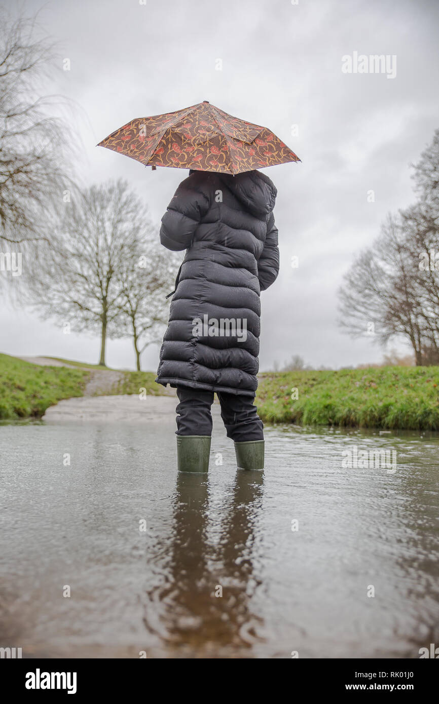 Kidderminster, Regno Unito. 8 febbraio, 2019. Regno Unito: meteo persistente pioggia pesante porta a livelli di acqua di risalita in Worcestershire con docce blustery continuando per tutto il giorno. Una giovane donna, vista posteriore, che indossa un lungo cappotto invernale e wellies sorge isolata all'esterno in acqua, lottando con il suo ombrello in umido e il vento. Credito: Lee Hudson/Alamy Live News Foto Stock