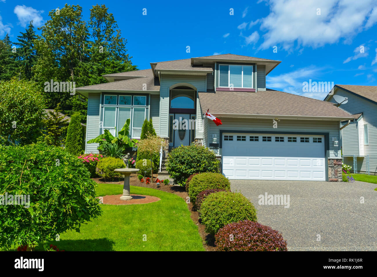 Casa residenziale con bandiera canadese all'ingresso e graziosamente decorato cortile anteriore sul cielo blu sullo sfondo. Casa Famiglia in British Columbia, Canada. Foto Stock