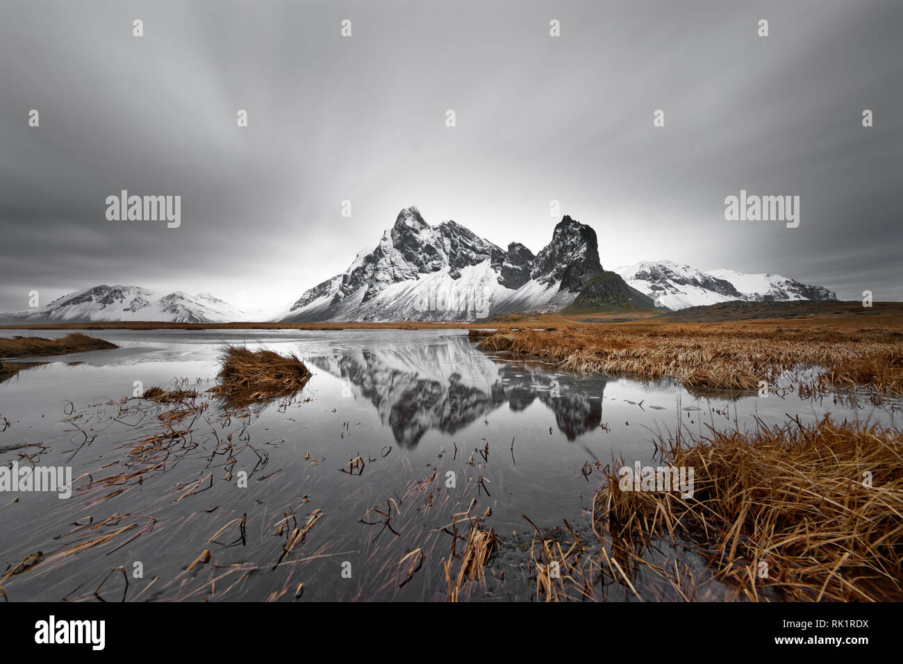 Coperto di neve e la formazione di montagna è riflessa in uno stagno, ciuffi di erba in e intorno allo stagno, lame di erba sono parzialmente spostata dal vento, cloud Foto Stock