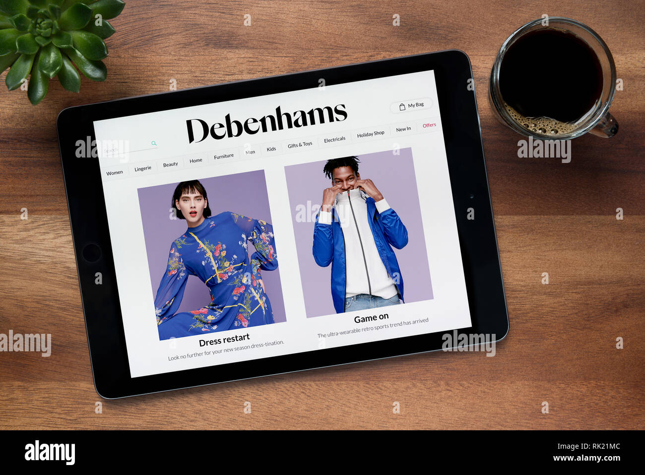 Il sito web di Debenhams è visto su un tablet iPad, su di un tavolo di legno lungo con un caffè espresso e una pianta di casa (solo uso editoriale). Foto Stock