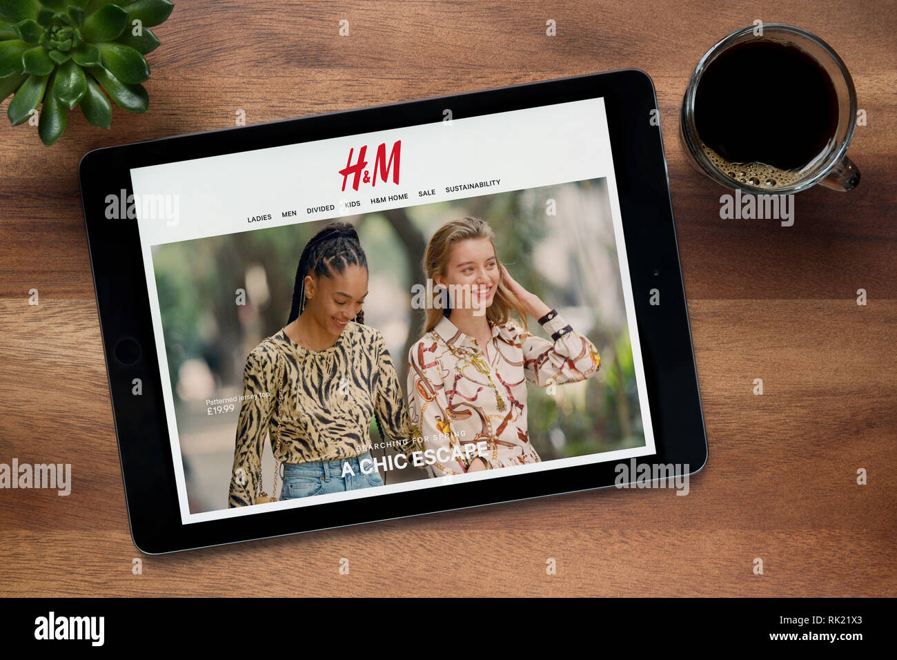 Il sito web di H&M è visto su un tablet iPad, su di un tavolo di legno lungo con un caffè espresso e una pianta di casa (solo uso editoriale). Foto Stock