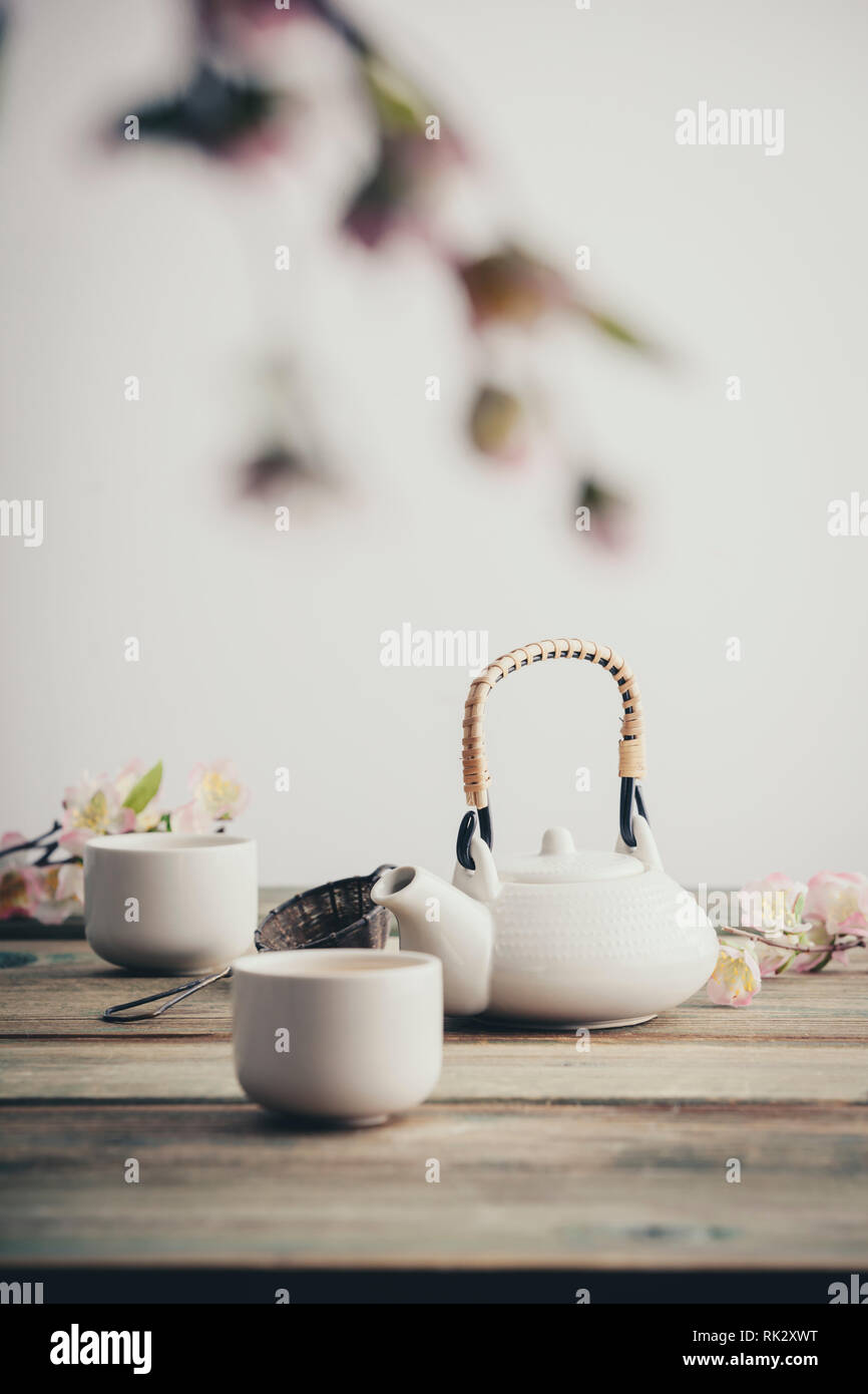 Tradizionali asiatici della cerimonia del tè disposizione. Teiera bianco, tazze, sakura fiori sul tavolo di legno contro il muro bianco, il fuoco selettivo, spazio di copia Foto Stock
