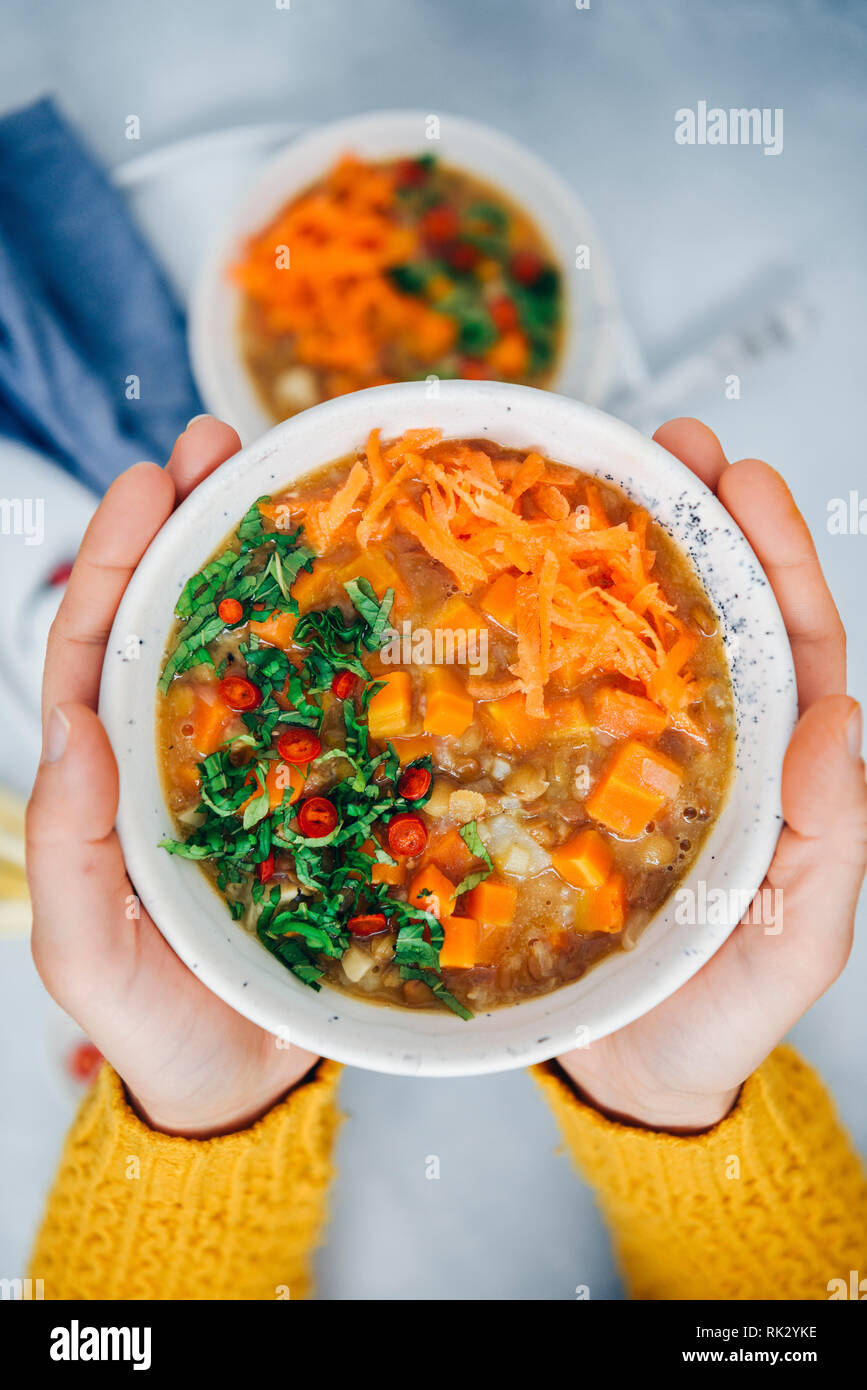 Una donna con giallo cardigan tenendo una ciotola di vegan zuppa di carote con riso e lenticchie e sedano rapa, guarnito con menta e peperoncino. Foto Stock