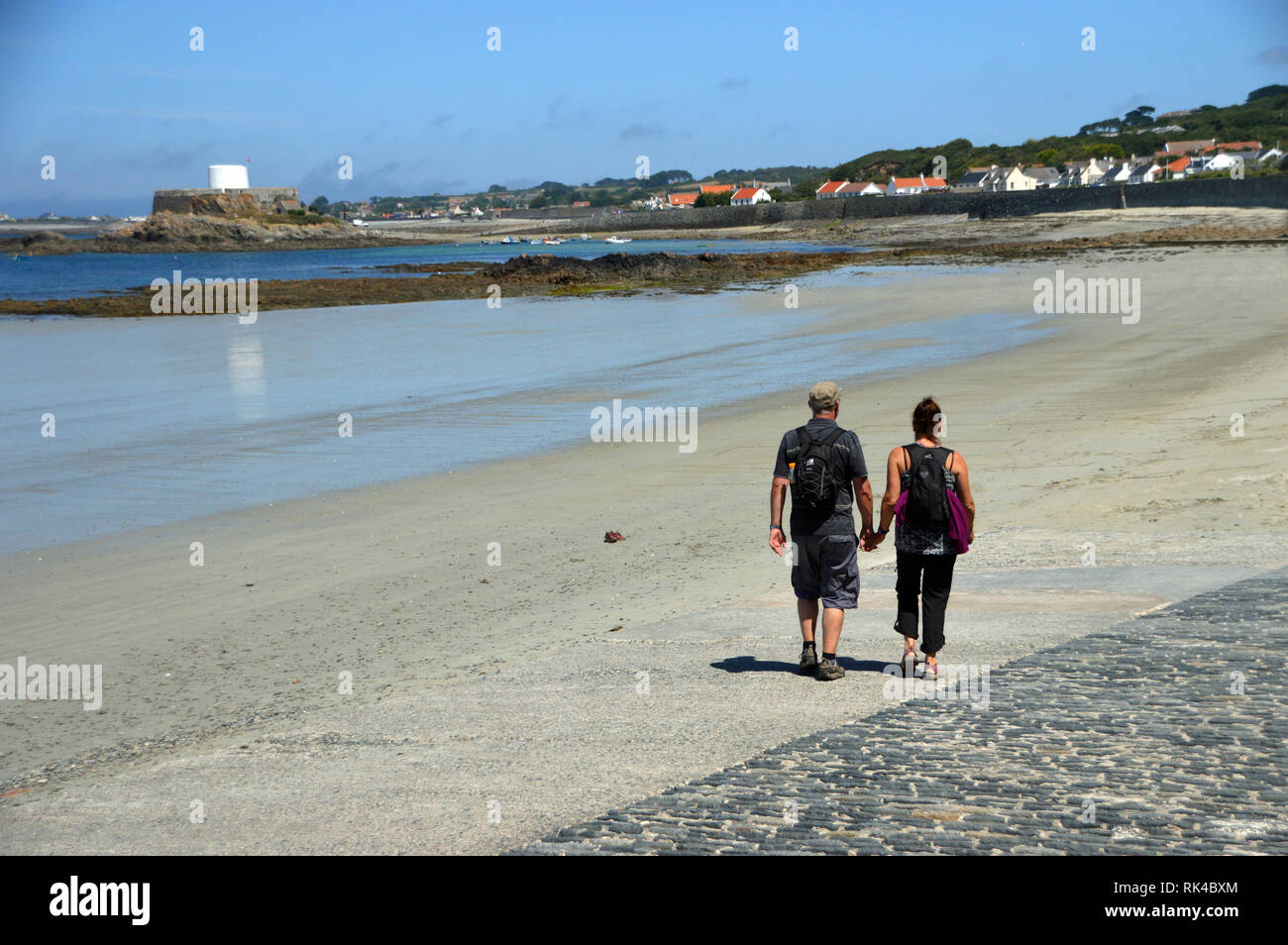 Giovane tenendo le mani e di camminare sulla spiaggia a Portelet Bay verso Fort Grey Shipwreck Museum 'martello' Torre in Guernsey, Isole del Canale.UK. Foto Stock