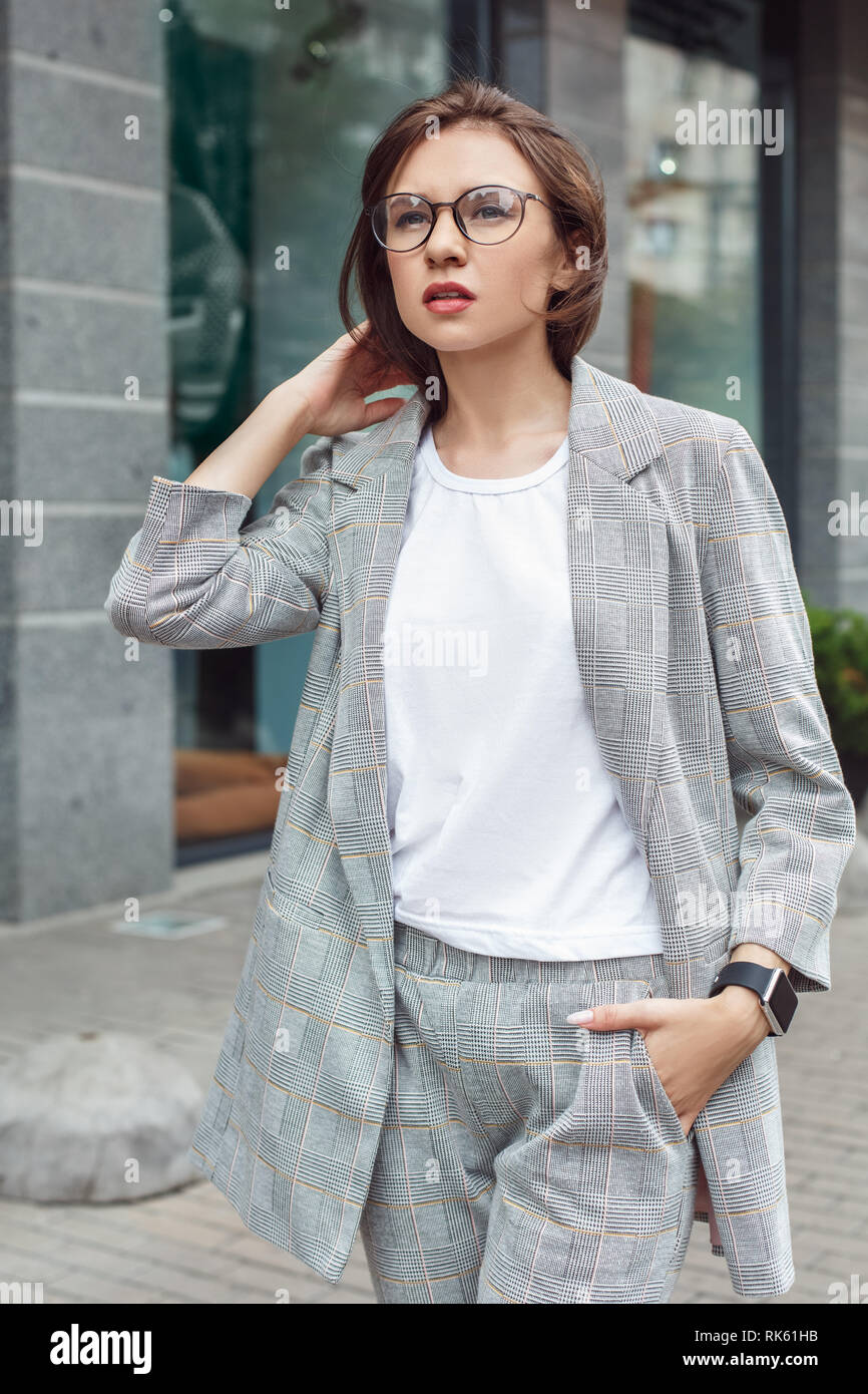 Business casual. Signora giovane in occhiali di camminare sulla via della città mano in tasca riflessivo Foto Stock