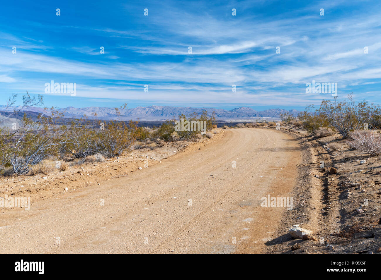 Deserto di strada sterrata che conduce verso le montagne al di là sotto il luminoso cielo blu con nuvole bianche. Foto Stock