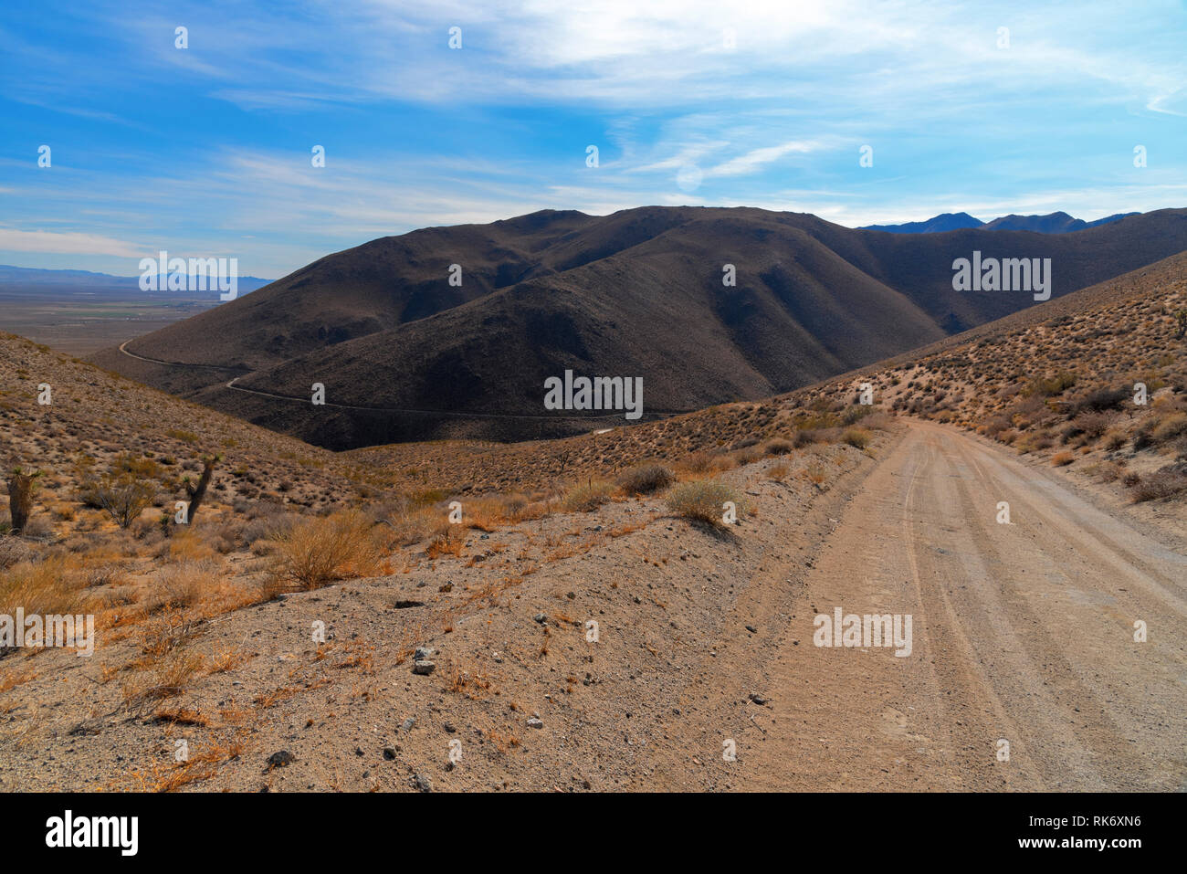 Deserto montagna strada sterrata che conduce verso la montagna sopra la valle sottostante sotto il luminoso cielo blu con nuvole bianche. Foto Stock