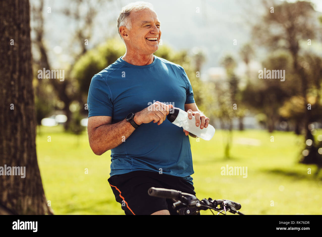 Senior uomo in usura fitness acqua potabile seduto su una bicicletta. Allegro senior persona fitness prendendo una pausa durante la pratica del ciclismo in un parco. Foto Stock