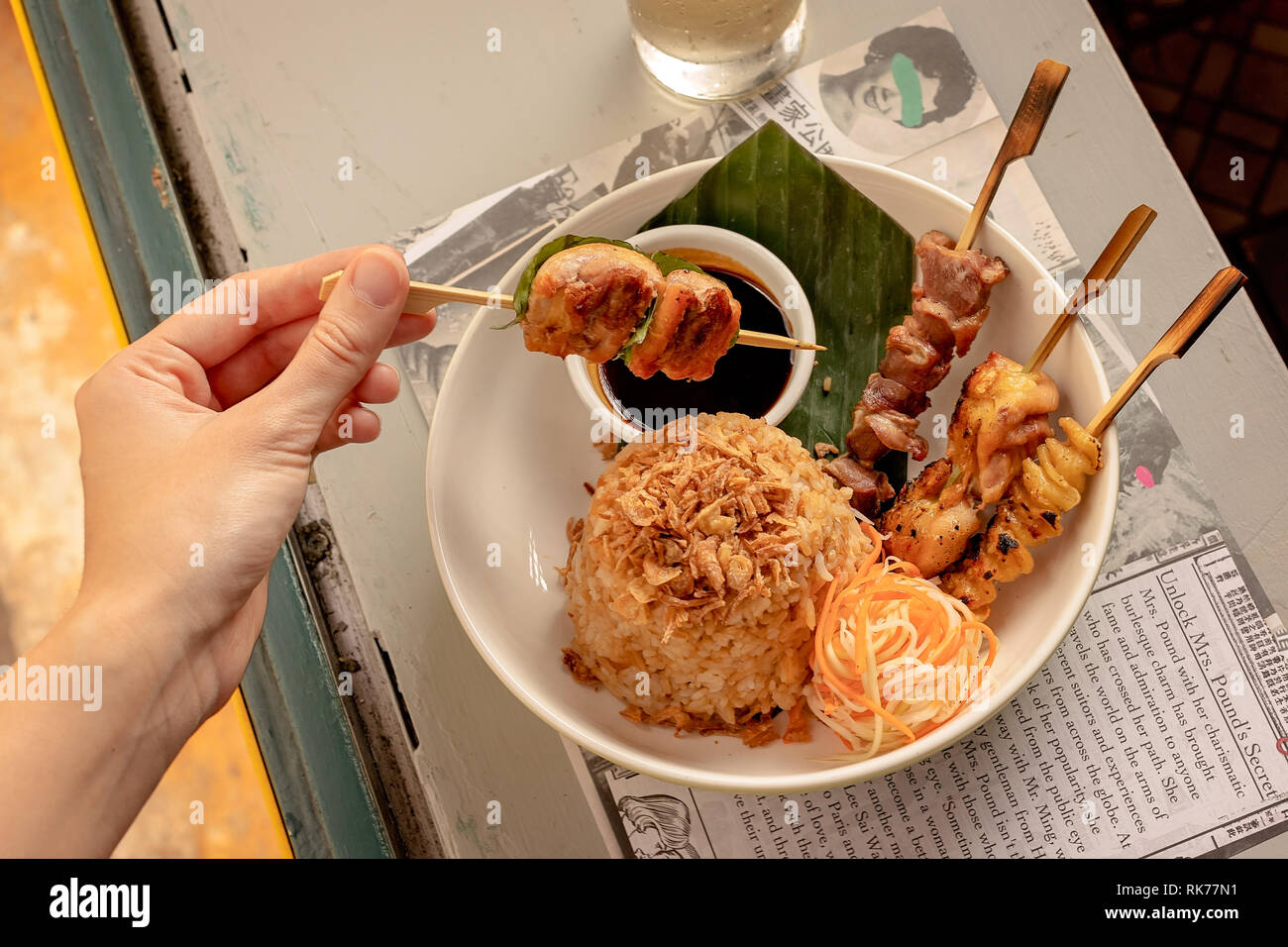 Una mano che tiene uno spiedino da una combinazione di carne servita con riso fritto, verdure e salsa di soia Foto Stock