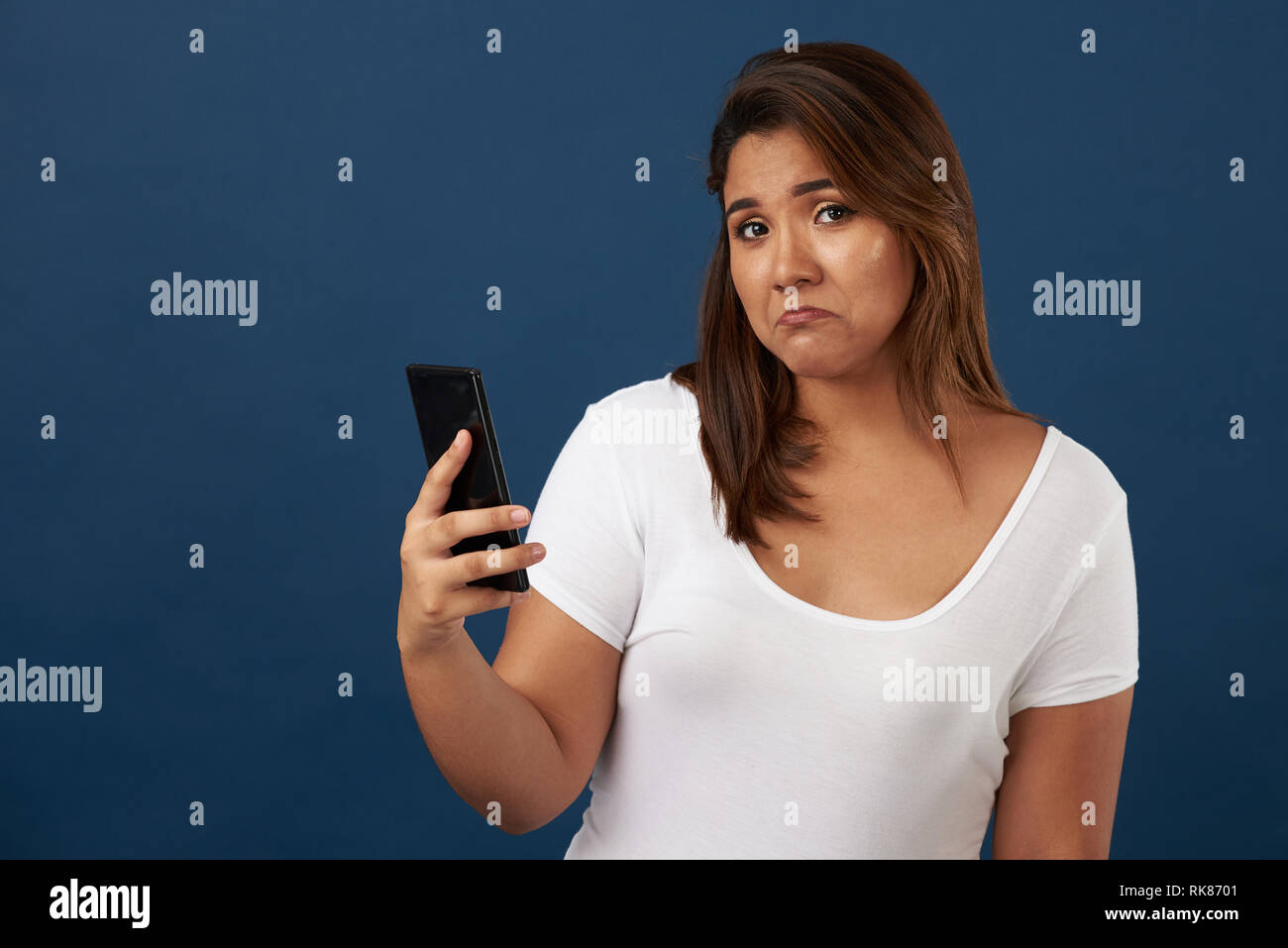 Sconvolto giovane donna con lo smartphone isolato su blu di sfondo per studio Foto Stock