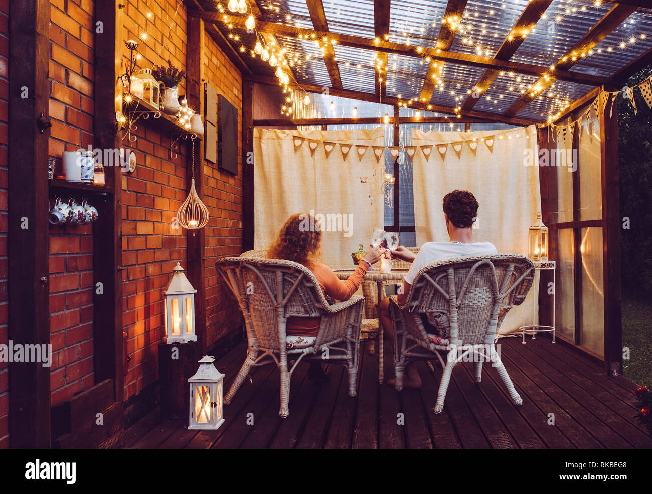 Coppia giovane uomo e donna sedersi su bianco rattan Sedie da giardino in legno sulla romantica terrazza all'aperto in serata la celebrazione e la tostatura, trascorrendo del tempo Foto Stock