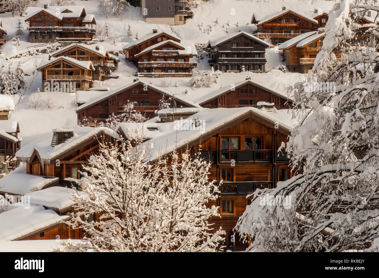 Dietro gli alberi, vista su un gruppo di coperta di neve la luce di legno marrone scuro e ski chalets in una località sciistica nelle Alpi francesi. Foto Stock