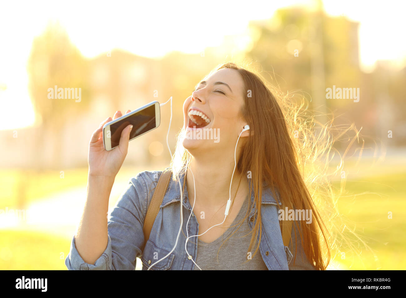 Felice ragazza cantare ascoltando la musica dal telefono intelligente in un parco al tramonto Foto Stock