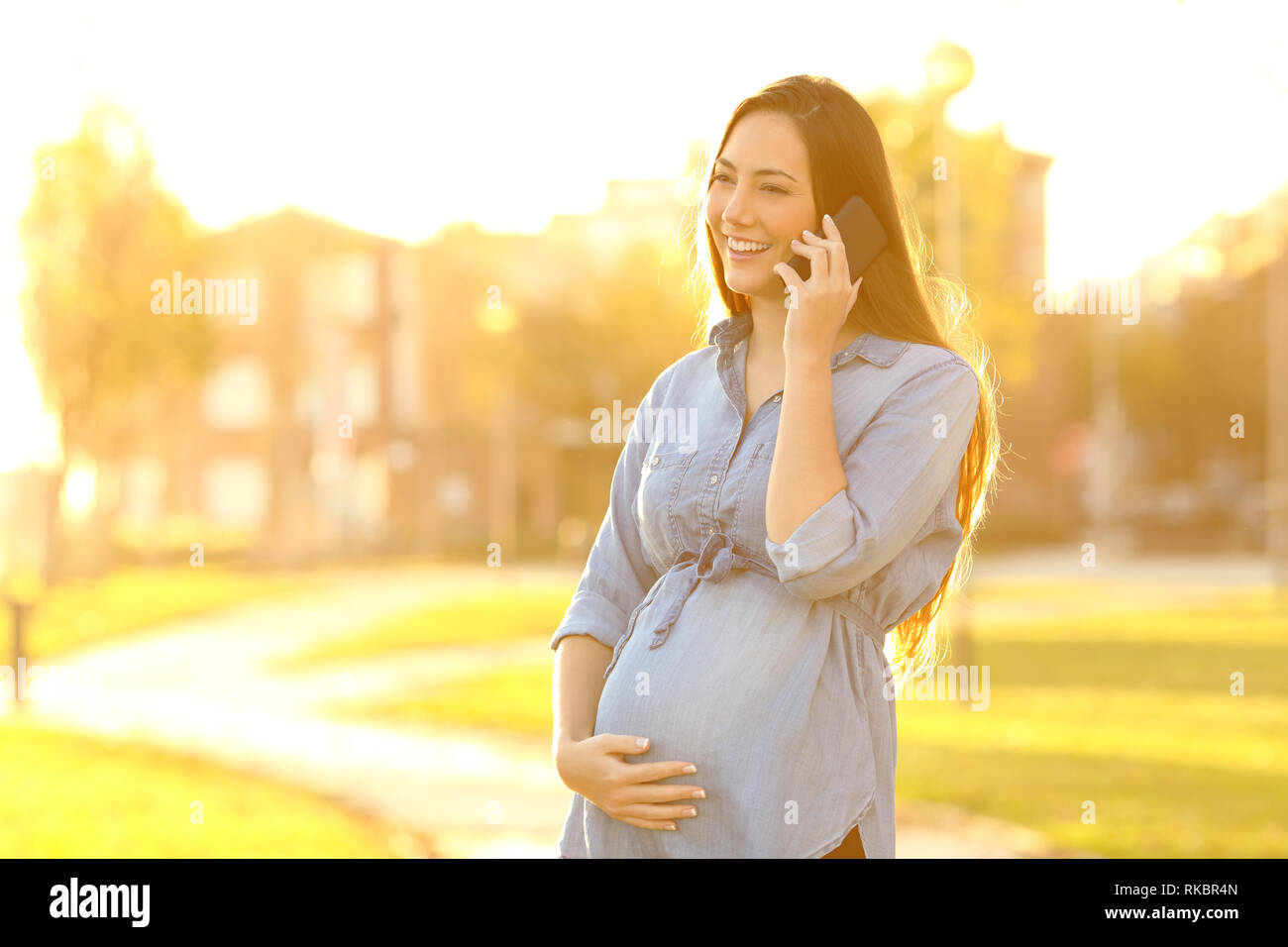 Felice donna incinta parlando al telefono in piedi al tramonto in un parco Foto Stock