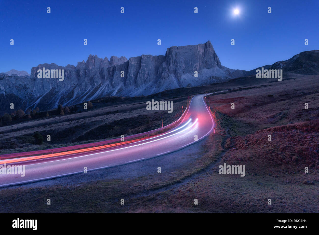 Sfocata fari auto sulla tortuosa strada di notte in autunno. Paesaggio con strada asfaltata, sentieri di luce, montagne, colline, blu cielo con i raggi della luna a dus Foto Stock