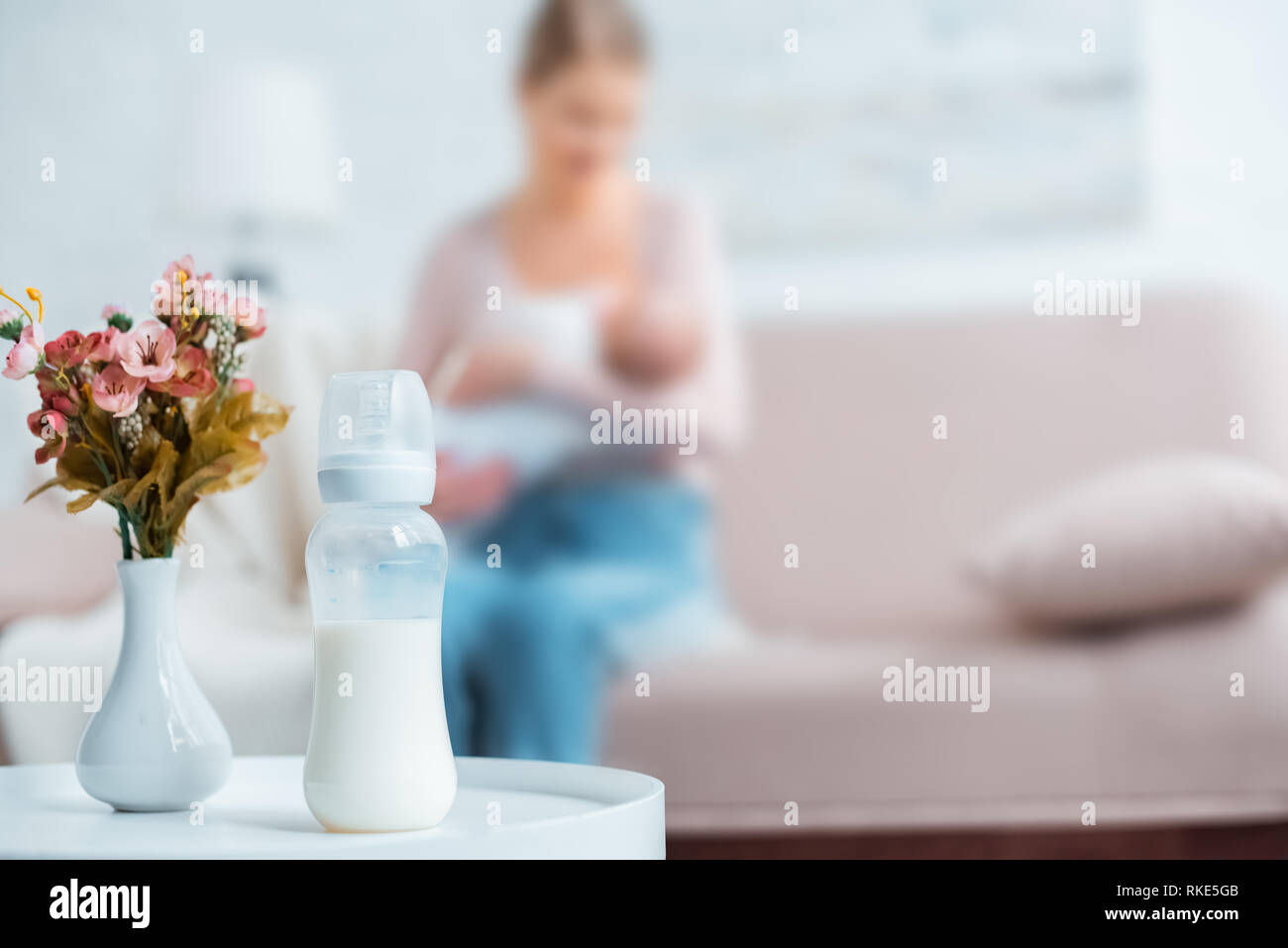 Vista ravvicinata di biberon con latte, fiori in vaso e madre di allattamento al seno baby dietro a casa Foto Stock
