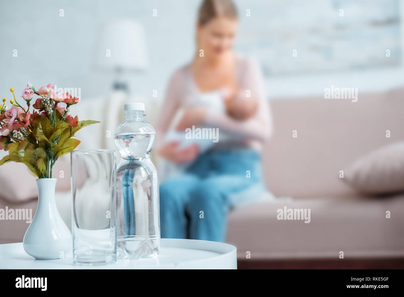 Vista ravvicinata di vetro, una bottiglia di acqua minerale, fiori in vaso e madre di allattamento al seno baby dietro a casa Foto Stock