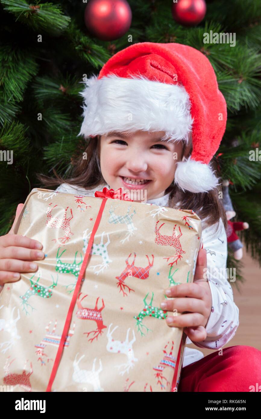 Gioiosa bambina bambino con la sua confezione regalo sotto albero di Natale. Lei ha appena hanno trovato il suo presente. Foto Stock