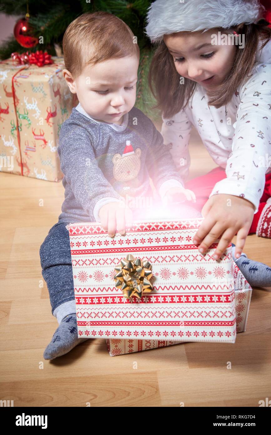 I piccoli fratelli di aprire le loro presenta vicino ad albero di Natale. Sono illuminate da luce magica dalla confezione regalo. Foto Stock