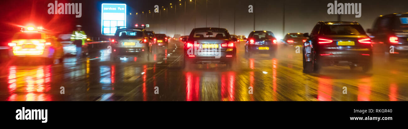 Panoramica banner web moto fotografia sfocata del traffico nella notte sotto la pioggia su una autostrada britannica con il funzionario di polizia e auto Foto Stock