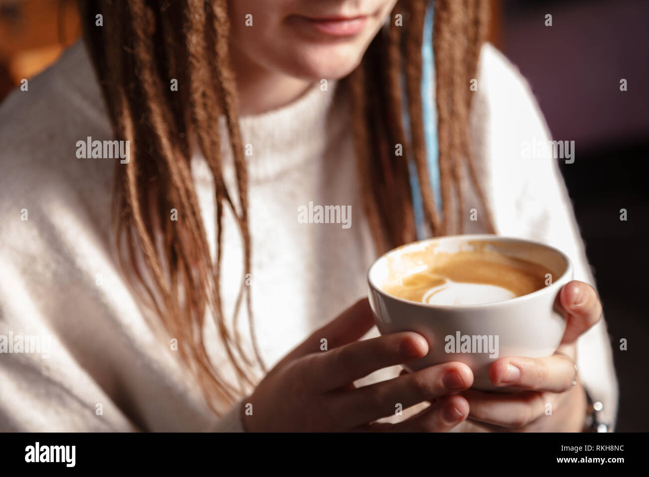 Un bianco di tazza di latte caldo art caffè con una forma di cuore nelle mani di una giovane ragazza con Dreadlocks. Donna che indossa caldo inverno a maglia maglione bianco. Foto Stock