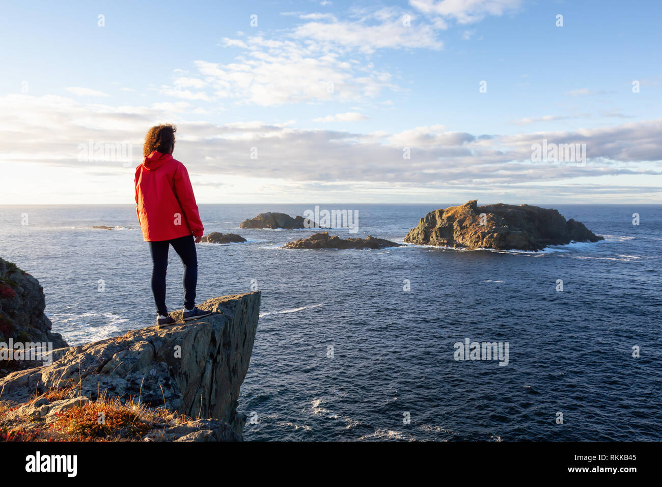 Woman in Red jacket è in piedi sul bordo di una scogliera e godere degli splendidi paesaggi dell'oceano. Preso in testa di corvo, Nord Twillingate Isola, Newfoun Foto Stock