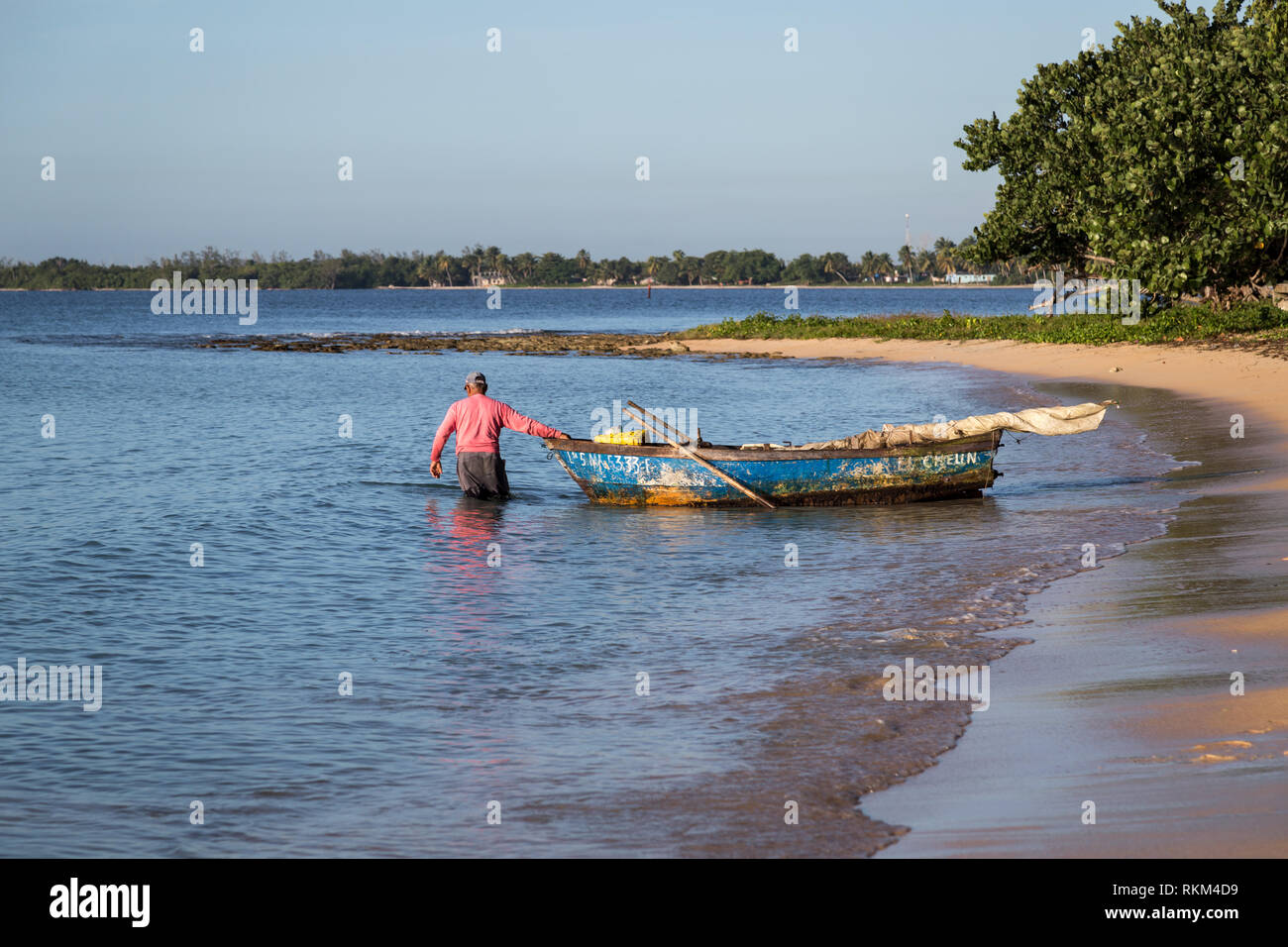 Pescatore cubano sta tirando fuori il suo vecchio blu barca con vele arrotolata in mattinata dalla spiaggia di sabbia nella speranza di catturare qualche pesce. Cuba Foto Stock