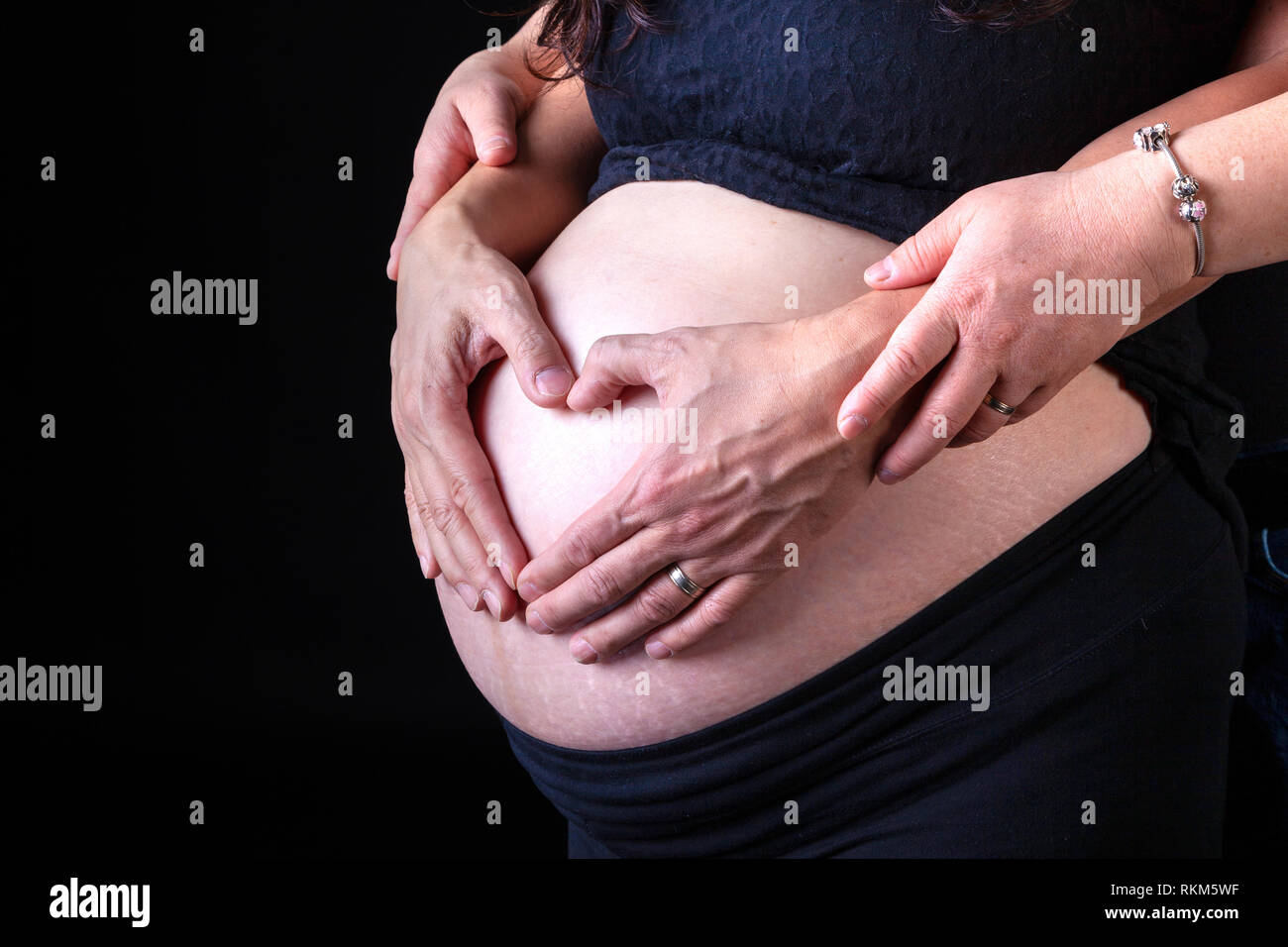 Donna incinta con il marito le mani sulla sua pancia nella forma di un cuore che simboleggia l'amore. Isolato su sfondo nero. Foto Stock