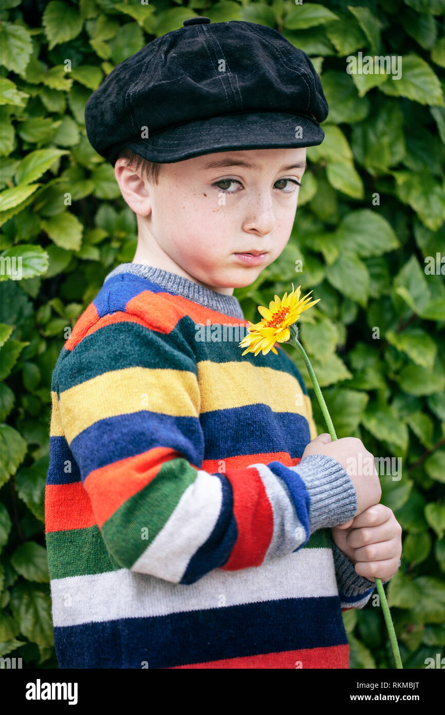 Triste, cute little boy in arcobaleno a strisce multicolori maglione e in camoscio nero newsboy cap tenendo un arancione fiore a margherita Foto Stock